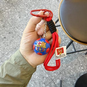 Puede incluir: Un llavero con un bal&oacute;n de f&uacute;tbol en miniatura de paneles azules y rojos con el escudo del FC Barcelona. Incluye un mosquet&oacute;n rojo, una secci&oacute;n de cord&oacute;n trenzado negro, una correa roja larga y una etiqueta blanca con el texto Replay y m&aacute;s texto.