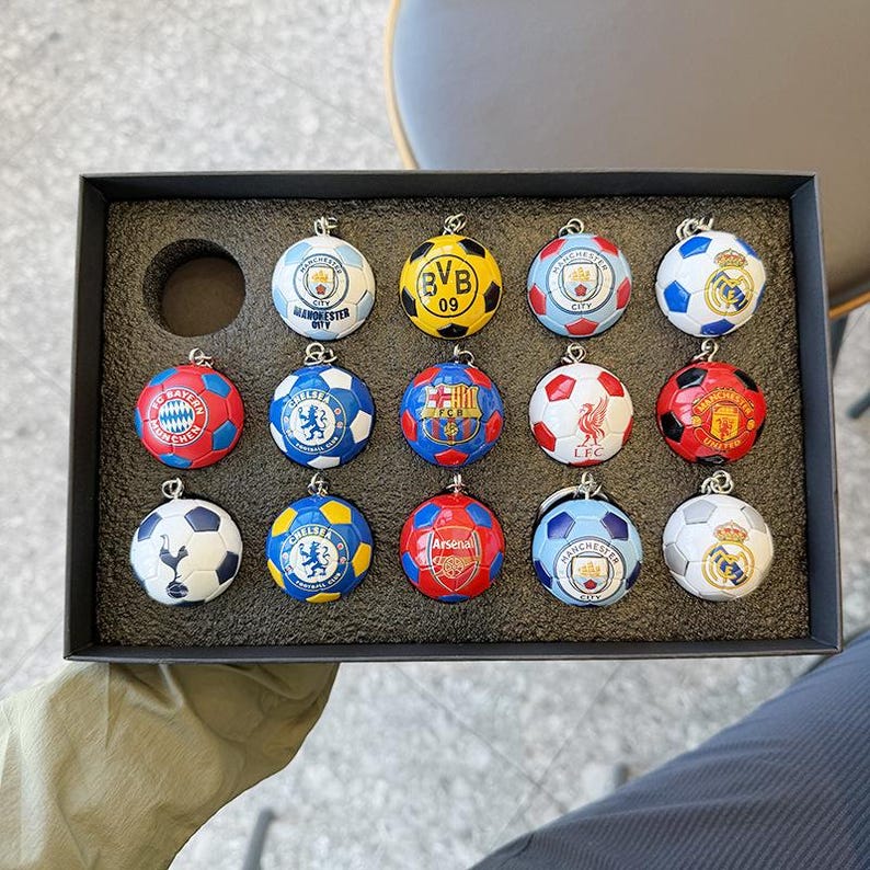 Puede incluir: Una caja de presentaci&oacute;n negra que contiene catorce llaveros esf&eacute;ricos con forma de bal&oacute;n de f&uacute;tbol. Cada llavero presenta el logotipo de un club de f&uacute;tbol profesional &uacute;nico, incluyendo Manchester City, Borussia Dortmund, Bayern Munich y Chelsea.