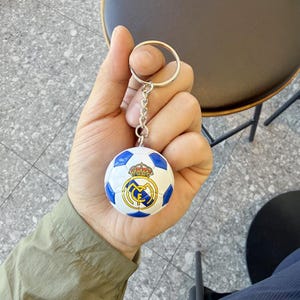 Puede incluir: Un llavero esf&eacute;rico con forma de bal&oacute;n de f&uacute;tbol que presenta un dise&ntilde;o de paneles blancos y azules con el logotipo del club Real Madrid impreso en el frente. El llavero incluye una anilla de metal plateado y una cadena corta.
