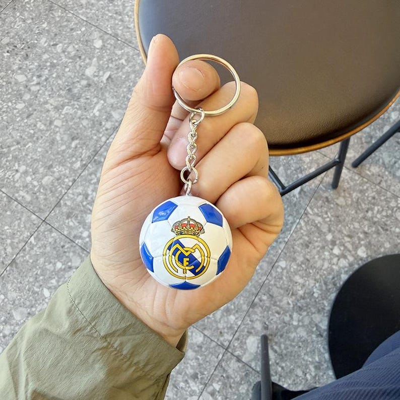 Puede incluir: Un llavero esf&eacute;rico con forma de bal&oacute;n de f&uacute;tbol que presenta un dise&ntilde;o de paneles blancos y azules con el logotipo del club Real Madrid impreso en el frente. El llavero incluye una anilla de metal plateado y una cadena corta.