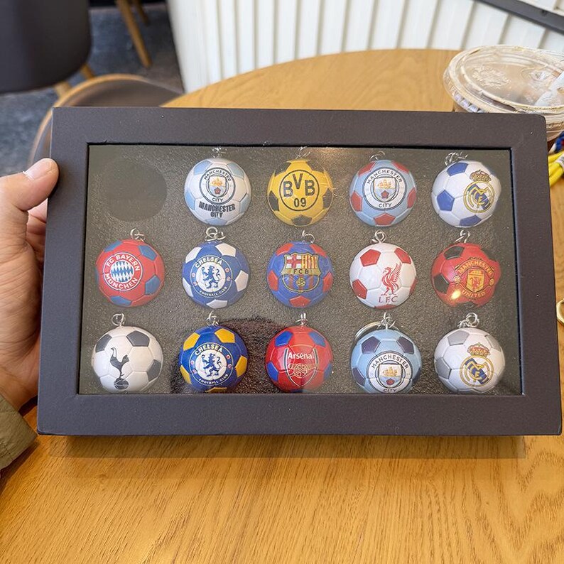 Puede incluir: Una caja expositora rectangular negra que contiene catorce peque&ntilde;os llaveros esf&eacute;ricos con forma de bal&oacute;n de f&uacute;tbol. Cada llavero presenta el logotipo de un club de f&uacute;tbol profesional diferente, incluidos Manchester City, BVB 09, FC Barcelona, Liverpool y Real Madrid.