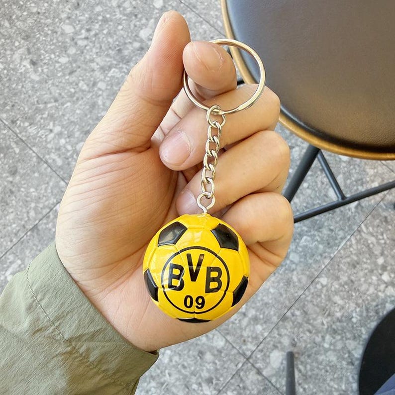 Puede incluir: Un llavero redondo con un dise&ntilde;o de bal&oacute;n de f&uacute;tbol en miniatura de color amarillo y negro. El bal&oacute;n muestra el texto negro BVB 09 en el centro. Est&aacute; unido a una cadena de metal plateado y una anilla circular para llaves.