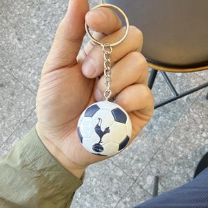 Puede incluir: Un llavero esf&eacute;rico de bal&oacute;n de f&uacute;tbol con un patr&oacute;n cl&aacute;sico de pent&aacute;gonos blancos y azul oscuro. El bal&oacute;n est&aacute; unido a una anilla de metal plateado mediante una cadena corta. Un emblema de gallo azul oscuro est&aacute; impreso en el bal&oacute;n.