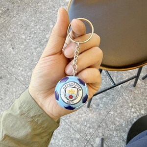 Puede incluir: Un llavero redondo con forma de bal&oacute;n de f&uacute;tbol que presenta el logotipo del club de f&uacute;tbol Manchester City en el centro. El bal&oacute;n tiene un dise&ntilde;o de paneles en azul claro y azul oscuro. Est&aacute; unido a una anilla de metal plateado por una cadena.