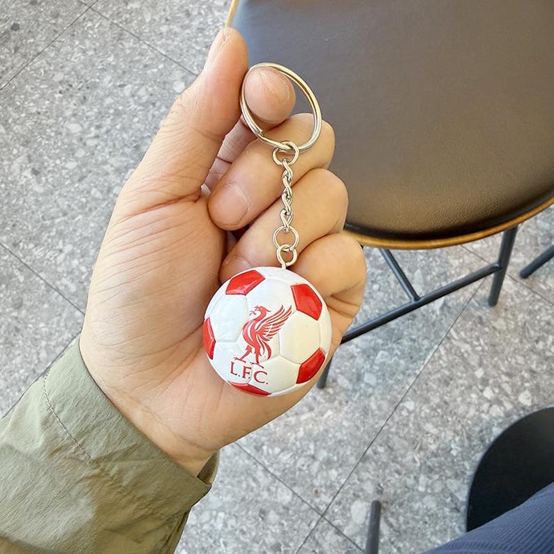 Puede incluir: Un llavero esf&eacute;rico de bal&oacute;n de f&uacute;tbol con superficie blanca y paneles pentagonales rojos. El centro del bal&oacute;n muestra un emblema rojo de un p&aacute;jaro Liver sobre el texto L.F.C. El bal&oacute;n est&aacute; unido a una cadena de metal plateado y una anilla.
