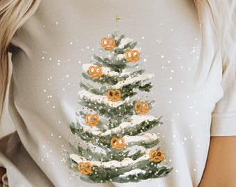 Könnte beinhalten: Cremefarbenes T-Shirt mit einer Aquarellillustration eines Weihnachtsbaums, der mit Brezeln geschmückt ist. Der Baum ist grün mit weißem Schnee und goldenen Akzenten. Das Shirt hat kurze Ärmel und einen Rundhalsausschnitt.