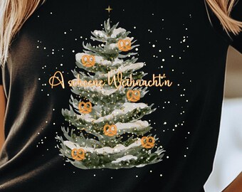 Könnte beinhalten: Schwarzes T-Shirt mit einem Weihnachtsbaum-Design, das mit Brezel-Ornamenten und dem Text "A schöne Weihnachtn" verziert ist. Der Baum hat goldene Akzente und einen Stern. Weiße Punkte sind um den Baum verstreut.