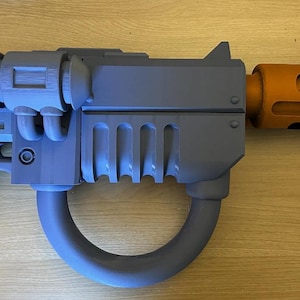 May include: A grey toy blaster with a prominent circular handle and a cylindrical orange barrel extension. The main body features a boxy design with a top-mounted component, a ribbed section, and a pistol-style grip, all rendered in a matte finish.