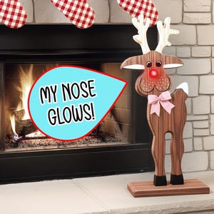 Puede incluir: Decoración navideña de reno de madera con nariz roja brillante y lazo rosa. El reno está sobre una base de madera frente a una chimenea con un bocadillo que dice "MY NOSE GLOWS!". Encima, cuelgan calcetines a cuadros rojos y blancos.
