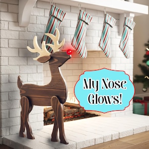 Puede incluir: Una decoración de reno de madera con una nariz roja brillante se encuentra frente a una chimenea. El reno tiene cuernos de color claro y está hecho de madera marrón oscura. El texto "My Nose Glows!" está en una burbuja azul. Cuelgan calcetines de Navidad.
