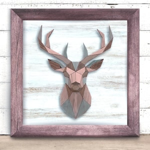 May include: Geometric wooden deer head art piece in a mauve-colored frame. The deer head is constructed from various shades of wood, including gray, brown, and pink. The background is a white-washed wood panel. The frame is a matching mauve color.