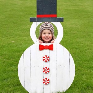 Puede incluir: Un recorte de muñeco de nieve de madera blanca con un sombrero de copa negro y una pajarita roja. Un niño con un sombrero sonríe a través de la abertura de la cara del muñeco de nieve. Caramelos de menta roja y blanca están en el cuerpo del muñeco de nieve.