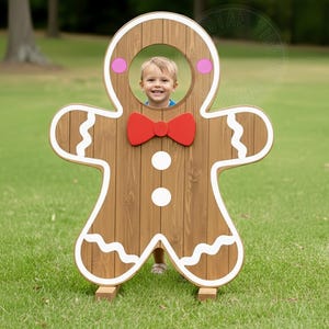 May include: A wooden gingerbread man photo prop with a child's face visible in the head cutout. The prop features a red bow tie, white icing details, and pink cheek accents. The gingerbread man stands on wooden supports.