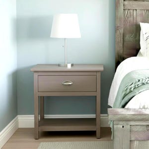 Puede incluir: Una mesita de noche de madera gris con un cajón y un estante. Una lámpara blanca con base plateada está encima. La mesita está junto a una estructura de cama de madera con un edredón blanco y una manta verde.