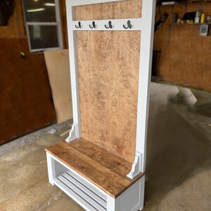 May include: A wooden entryway bench with a white frame and a natural wood seat and back panel. The bench features four black hooks and a lower shelf for storage. The design is a combination of rustic and modern styles.