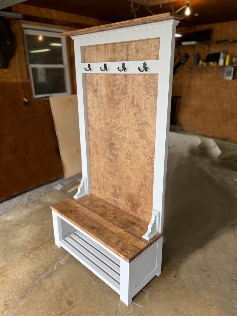 May include: A wooden entryway bench with a white frame and a natural wood seat and back panel. The bench features four black hooks and a lower shelf for storage. The design is a combination of rustic and modern styles.