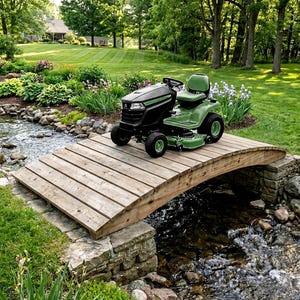 May include: A black and green riding lawn mower with a steering wheel, a padded seat, and a cutting deck, positioned on a curved wooden footbridge that spans a small rocky stream in a garden setting with green grass and various plants.