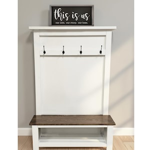 May include: A white entryway bench with a dark wood seat and a white backboard. The backboard has four black hooks and a framed sign that reads "this is us" in white script.