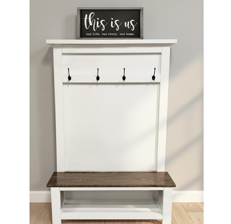 May include: A white entryway bench with a dark wood seat and a white backboard. The backboard has four black hooks and a framed sign that reads "this is us" in white script.