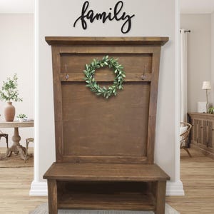 May include: A wooden entryway bench with a back panel and a lower shelf. The bench is stained a medium brown and features two hooks and a green wreath. Above the bench, the word "family" is displayed in black script.