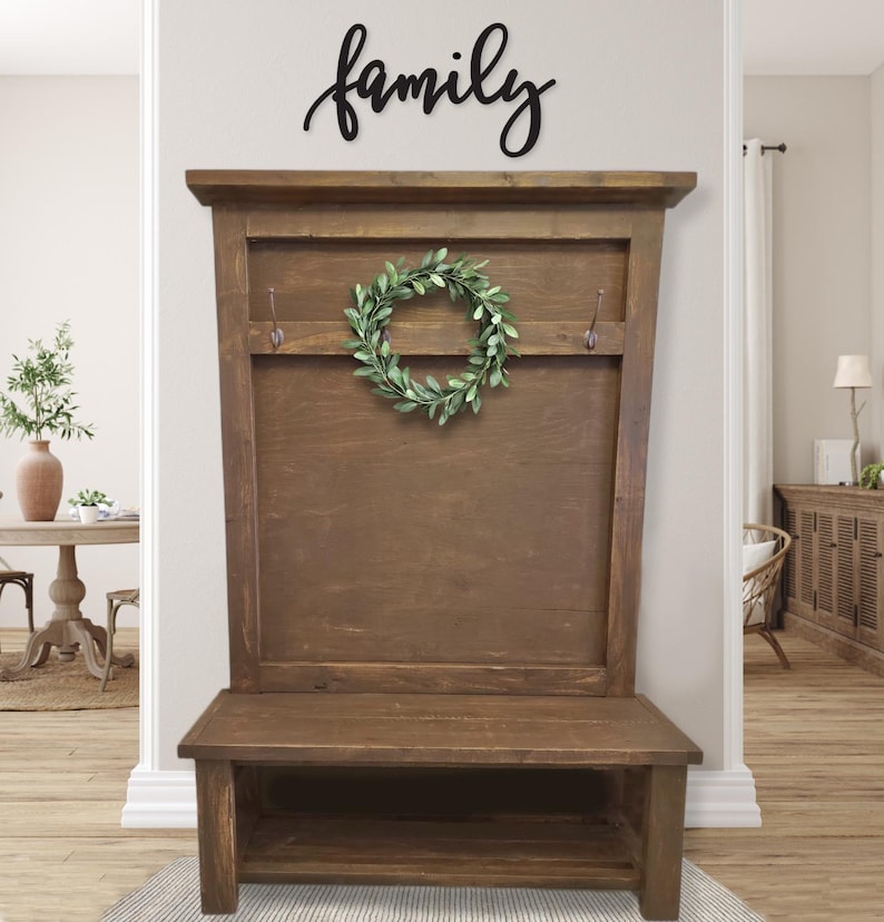 May include: A wooden entryway bench with a back panel and a lower shelf. The bench is stained a medium brown and features two hooks and a green wreath. Above the bench, the word "family" is displayed in black script.