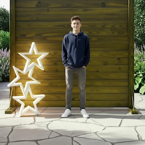May include: A young person stands in front of a wooden fence, with a set of three white star-shaped lights. The person is wearing a navy blue hoodie, gray pants, and white sneakers. The lights are on a stone patio.