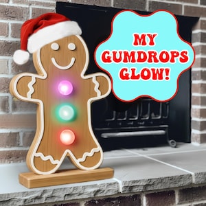 May include: A wooden gingerbread man Christmas decoration wearing a Santa hat. The gingerbread man has three glowing gumdrop lights in pink, green, and red. The text "MY GUMDROPS GLOW!" is in a speech bubble.