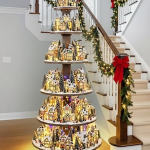 May include: A multi-tiered Christmas village display with illuminated miniature houses and trees. The display is on a wooden stand and is decorated with snow. The text at the bottom reads "DISASSEMBLES & REASSEMBLES EASILY!"