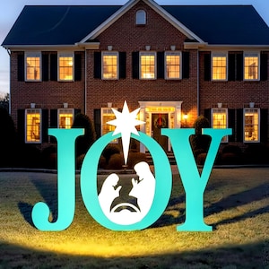 May include: A teal-colored outdoor Christmas decoration spelling out the word "JOY" with a nativity scene inside the "O". A large white star is above the "O". The decoration is in front of a two-story brick house with lit windows.