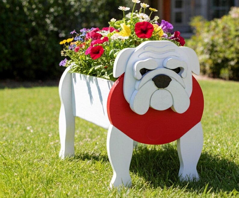 K&ouml;nnte beinhalten: Ein wei&szlig;er Holzblumentopf in Form einer Bulldogge mit rotem Halsband und schwarzer Nase. Der Topf ist mit bunten Blumen gef&uuml;llt, darunter rote, gelbe und violette Bl&uuml;ten. Der Topf steht auf einem gr&uuml;nen Rasen.