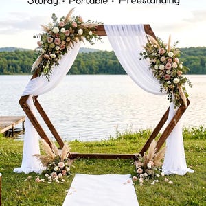 May include: A hexagonal wooden arch featuring the text Sturdy • Portable • Freestanding. The dark brown frame is draped with white fabric and decorated with floral arrangements of pink roses, white flowers, and pampas grass on the top and base.