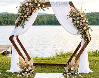 May include: A hexagonal wooden arch featuring the text Sturdy • Portable • Freestanding. The dark brown frame is draped with white fabric and decorated with floral arrangements of pink roses, white flowers, and pampas grass on the top and base.