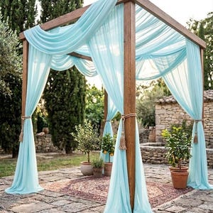 May include: A wooden pergola draped with flowing, light blue fabric. The structure has a square frame and is decorated with tassels. Potted plants and a patterned rug are visible beneath the pergola, creating an outdoor event space.