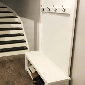 May include: A white wooden entryway bench with a back panel featuring four black hooks. The bench has a lower shelf and a storage compartment. The background includes a staircase and wood flooring.