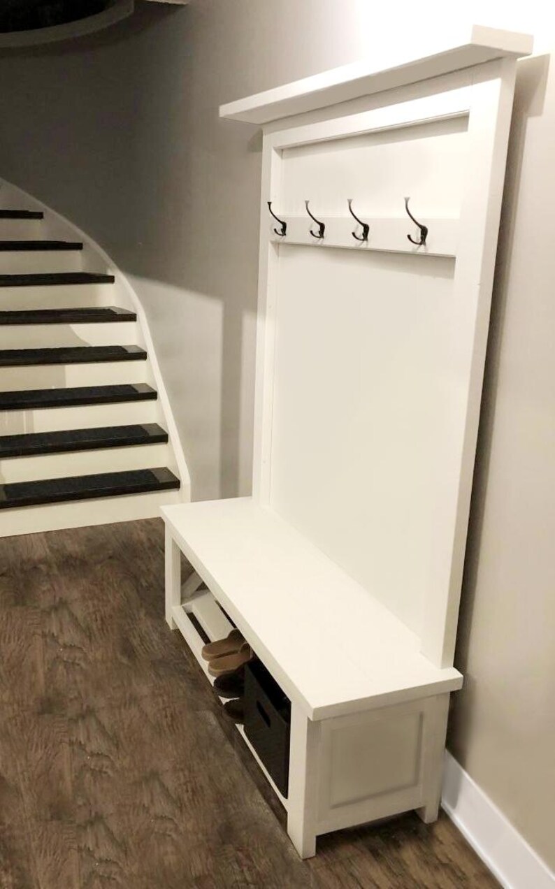 May include: A white wooden entryway bench with a back panel featuring four black hooks. The bench has a lower shelf and a storage compartment. The background includes a staircase and wood flooring.