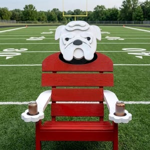 Op de afbeelding: Rode houten stoel met een bulldogkop en armen, met twee blikjes. De stoel staat op een voetbalveld. De bulldogkop is wit met zwarte accenten. De armen zijn wit met een bloemvormige bekerhouder.
