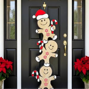 May include: A wooden Christmas door decoration featuring three stacked gingerbread men. The top gingerbread man wears a Santa hat and holds a candy cane. Each gingerbread man has a smiling face and snowflake accents.