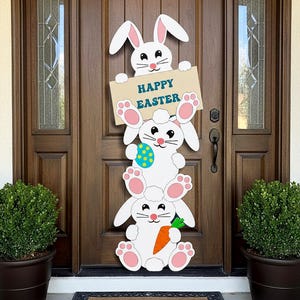 Op de afbeelding: Houten paasdeurdecoratie met drie gestapelde witte konijnen. Het bovenste konijn houdt een bord met de tekst "HAPPY EASTER" vast. Het middelste konijn houdt een blauw en geel ei vast en het onderste konijn een oranje wortel.