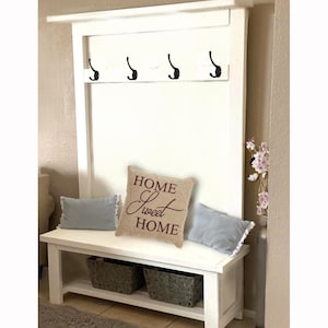 May include: A white entryway bench with a built-in coat rack. The bench has a lower shelf with two gray woven baskets. A decorative pillow with "HOME Sweet HOME" text sits on the bench, along with two light blue pillows.