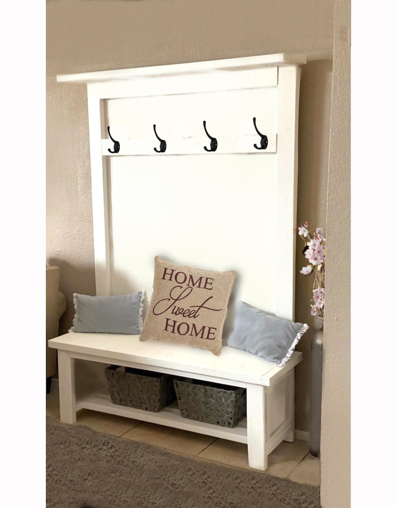 May include: A white entryway bench with a built-in coat rack. The bench has a lower shelf with two gray woven baskets. A decorative pillow with "HOME Sweet HOME" text sits on the bench, along with two light blue pillows.