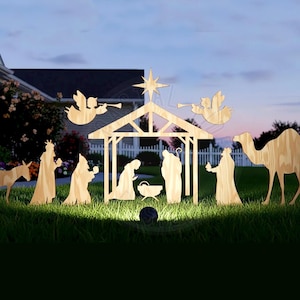 Peut inclure: Scène de la Nativité extérieure en bois, avec des silhouettes de la Sainte Famille, des rois mages, des anges, un âne et un chameau. Les personnages se détachent sur un ciel nocturne et de l'herbe verte.