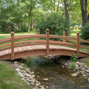 Op de afbeelding: Een houten tuinbrug met een gebogen ontwerp overspant een kleine beek. De brug is gemaakt van lichtbruin hout en heeft een lattenbodem en decoratieve balustrades. De achtergrond omvat weelderig groen gras, bomen en gebladerte.