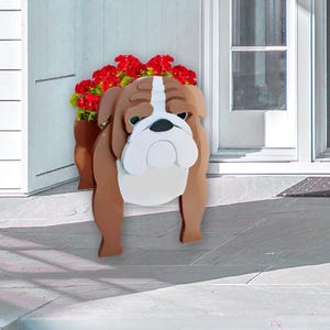 K&ouml;nnte beinhalten: Ein Holz-Blumentopf in Form einer Bulldogge, braun und wei&szlig;, gef&uuml;llt mit roten Blumen. Die Bulldogge hat einen traurigen Gesichtsausdruck und steht in der N&auml;he einer wei&szlig;en T&uuml;r.
