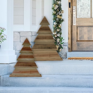May include: Two wooden Christmas tree decorations. The trees are made of horizontal planks of wood, creating a layered effect. The larger tree stands approximately 4 feet tall, while the smaller one is about 2 feet tall. They are placed on a porch.