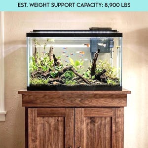 May include: A 25-gallon aquarium sits atop a solid wood stand. The aquarium is filled with plants, driftwood, and colorful fish. The stand has two doors with dark hardware and a weight support capacity of 8,900 lbs.