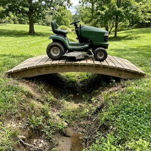 May include: A green riding lawn mower is parked on a small wooden bridge over a stream. The mower has a black seat and steering wheel. The bridge is made of light brown wood and is surrounded by green grass and trees.