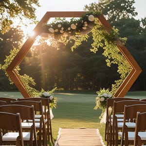 May include: A wooden hexagonal wedding arch decorated with greenery and white flowers, set up on a grassy lawn. Wooden chairs with white cushions are arranged in rows, leading to the arch. The sun shines brightly through the trees in the background.