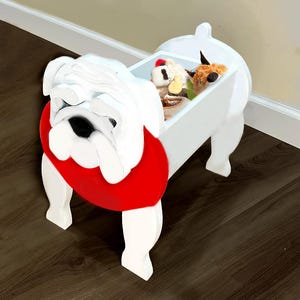 K&ouml;nnte beinhalten: Eine wei&szlig;e Holz-Aufbewahrungsbox in Form einer Bulldogge mit rotem Kragen. Die Box hat ein rechteckiges Staufach, das mit Stofftieren gef&uuml;llt ist. Die Bulldogge hat ein detailliertes Gesicht und steht auf vier Beinen.