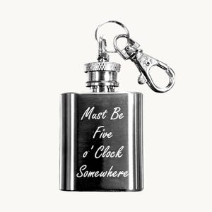 May include: A silver-toned metal flask keyring featuring a screw-top cap and a lobster claw clasp. The front of the rectangular flask is engraved with the white text "Must Be Five o' Clock Somewhere" in a cursive script font against the metallic surface.