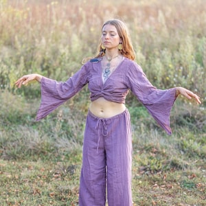 K&ouml;nnte beinhalten: Eine Frau tr&auml;gt ein lilafarbenes, lang&auml;rmeliges, locker sitzendes Oberteil mit einem geknoteten Vorderteil und eine passende weite Hose. Sie steht in einem Feld mit hohem Gras und hat die Arme ausgestreckt.